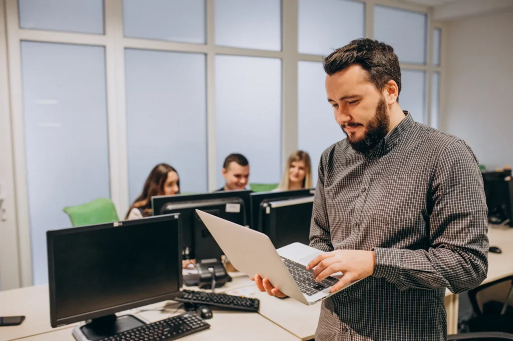Tecnico informatico in piedi con un portatile aperto in un ufficio con diverse postazioni di lavoro